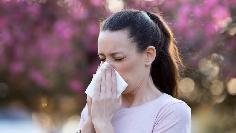 Woman blowing her nose outside with background flowers.